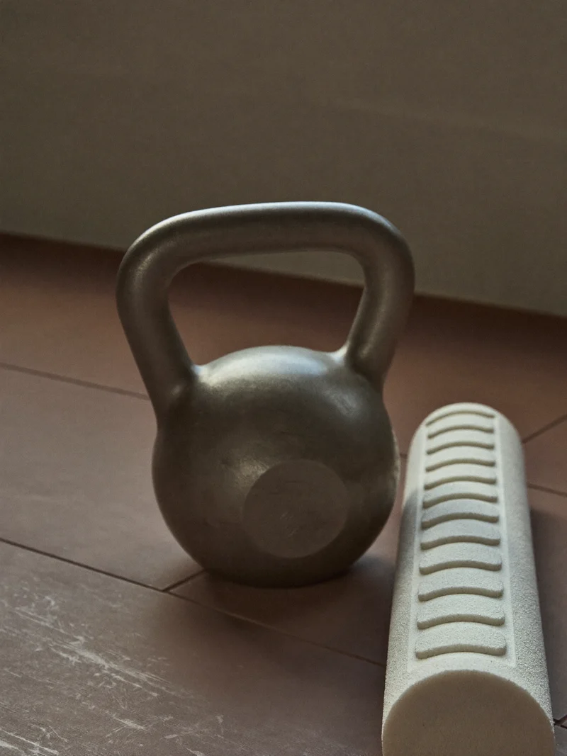 Mattierte Kettlebell neben einer Faszienrolle auf tonfarbenem Boden im diffusen Licht